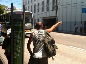 Homem com bolsa nas costa com o braço esticado solicitando para do ônibus que está chegando